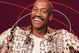 A smiling man with a bald head and mustache wears a patterned shirt against a maroon background with red and light blue curved lines.